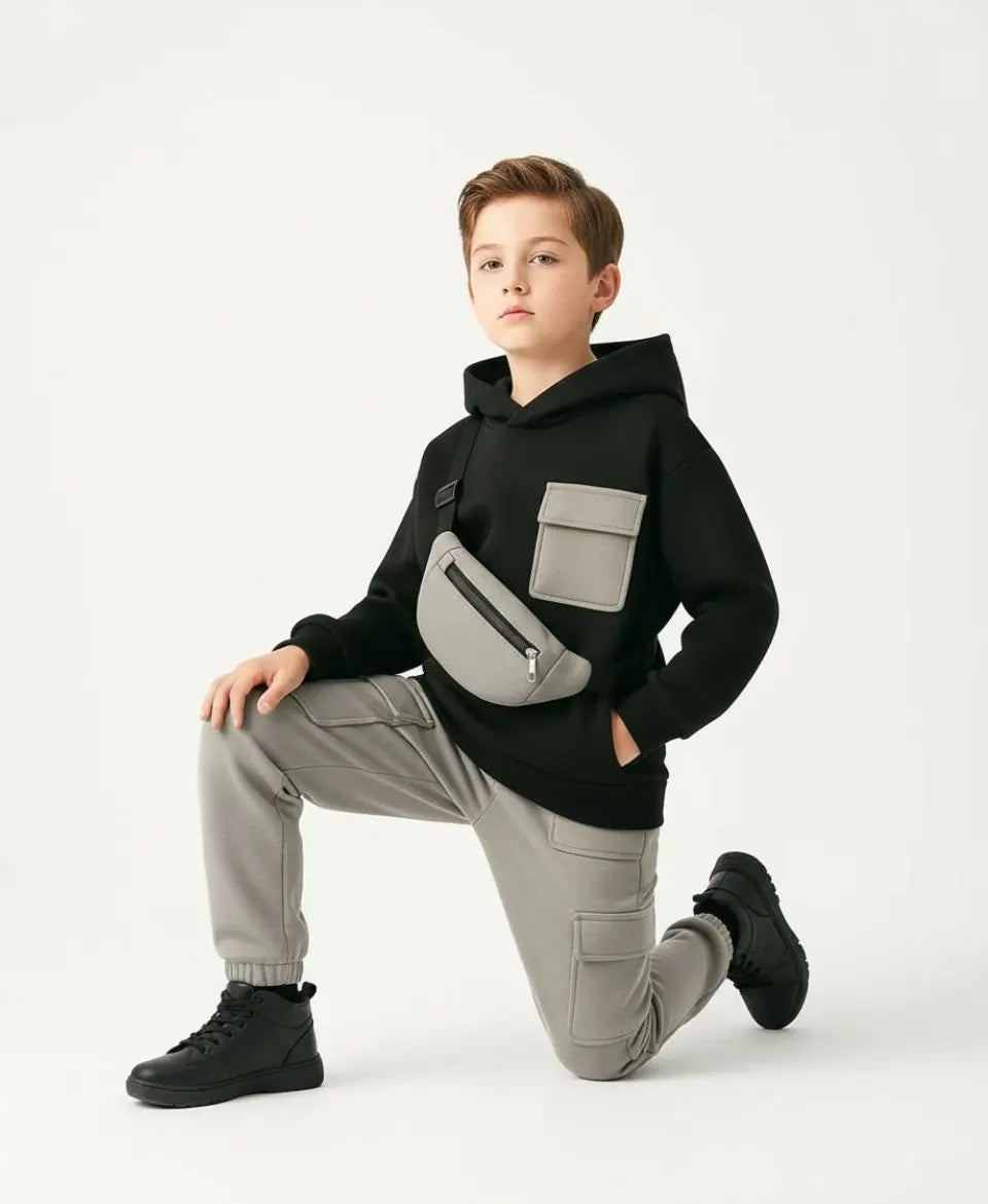 Child wearing a black hoodie with a gray bag, gray pants, and black shoes on a white background