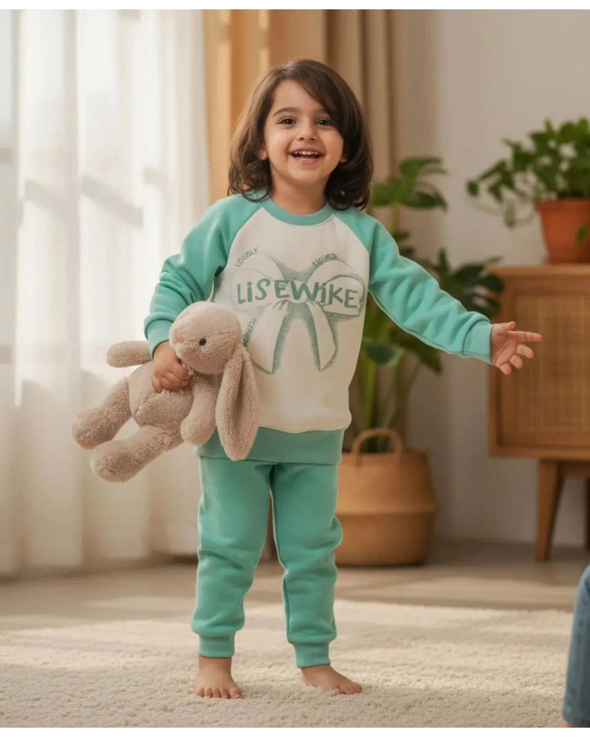 Child wearing a teal and white pajama set with a bunny design, holding a plush toy in a room with plants and a wooden cabinet.