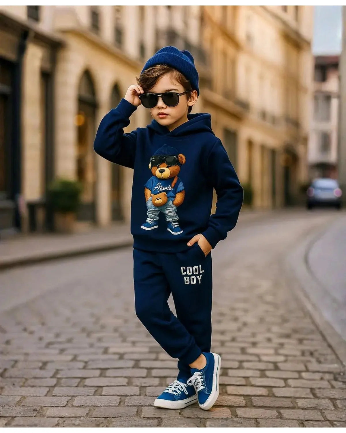 Happy Kidi Child wearing a navy blue outfit with a teddy bear graphic and 'Cool Boy' text, standing on a street.
