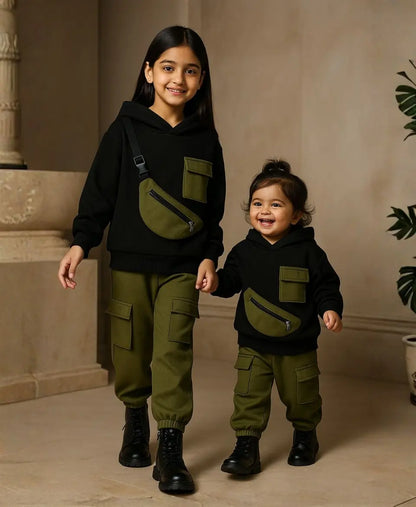 Happy Kidi Two children wearing matching black hoodies and green pants with fanny packs, standing indoors.