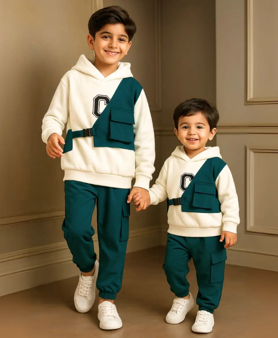 Two children wearing matching outfits with white hoodies and green pants, standing against a neutral wall.