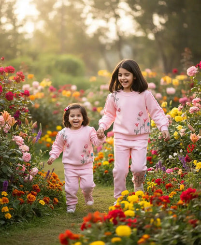 Two young girls in matching pink outfits standing in a garden filled with colorful flowers.